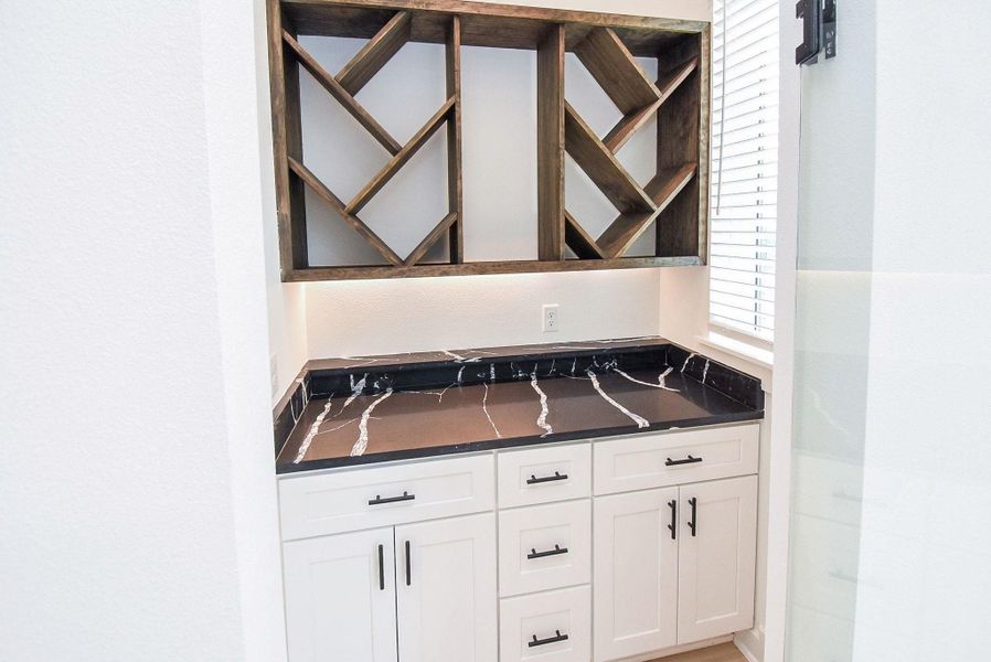 This space features a stylish wine rack with dark wood finish above a sleek black marble countertop. The white cabinetry offers ample storage, and the area is well-lit by natural light from a nearby window, creating an elegant and functional nook.