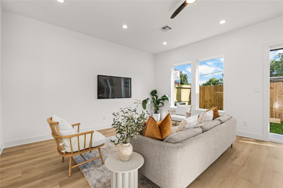 This view shows the spacious layout and natural light of the main living room. The light oak flooring easily fits a sofa and wood accent chairs, while large windows look out into the private backyard. A wall-mounted TV demonstrates the room's setup for entertainment.