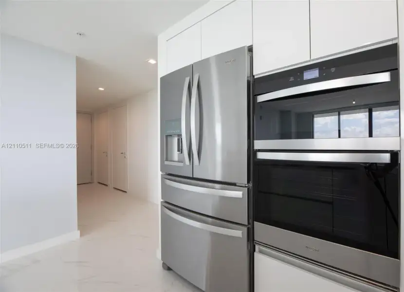 Stainless steel French door refrigerator and stacked double oven, complemented by white flat-panel cabinetry and large-format tile flooring