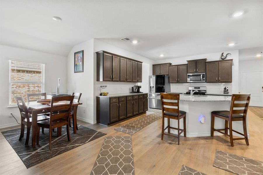 Kitchen with dark brown cabinetry, light stone counters, light wood finished floors, stainless steel appliances, and tasteful backsplash