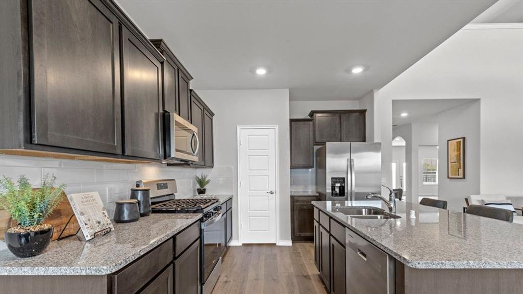 Kitchen featuring dark wood-finish cabinetry, light granite countertops, stainless steel appliances, a white subway tile backsplash, and wood-finish flooring Kitchen featuring dark wood-finish cabinetry, light granite countertops, stainless steel appliances, a white subway tile backsplash, and wood-finish flooring