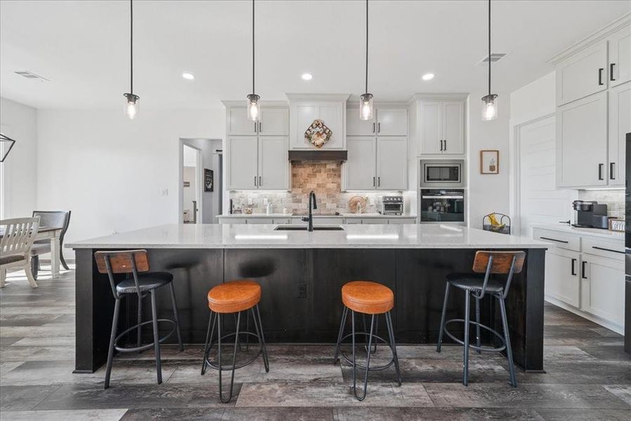 Kitchen with hanging light fixtures, backsplash, a breakfast bar, white cabinetry, and a large island with sink