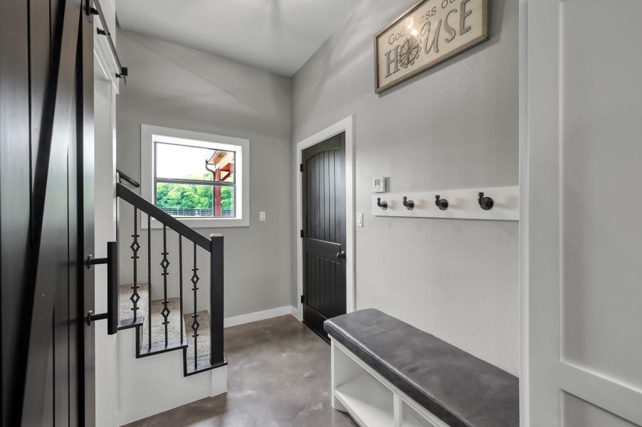 This entryway features modern design with a sleek black door, polished concrete floors, and a staircase with decorative iron railings. It includes a built-in bench with storage, wall hooks for convenience, and a window providing natural light.