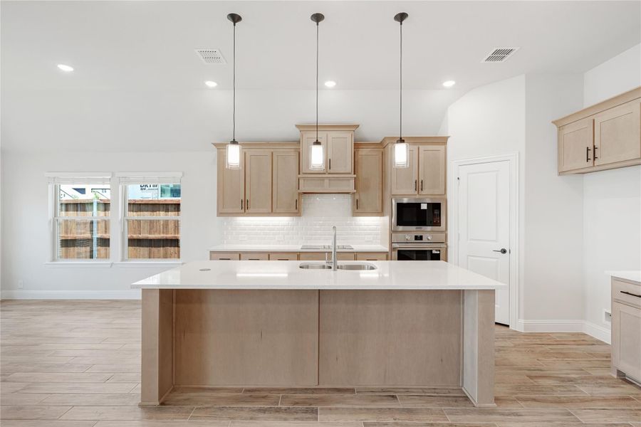 Kitchen with large island, light wood cabinets, white subway tile backsplash, and three pendant lights overhead.