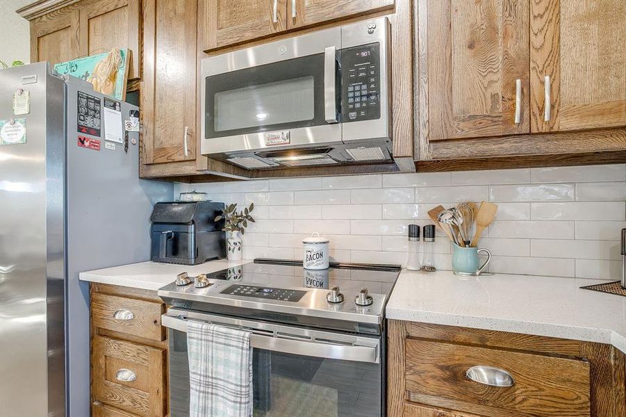 Kitchen with stainless steel appliances, tasteful backsplash, light quartz countertops, and brown cabinetry Kitchen with stainless steel appliances, tasteful backsplash, light quartz countertops, and brown cabinetry