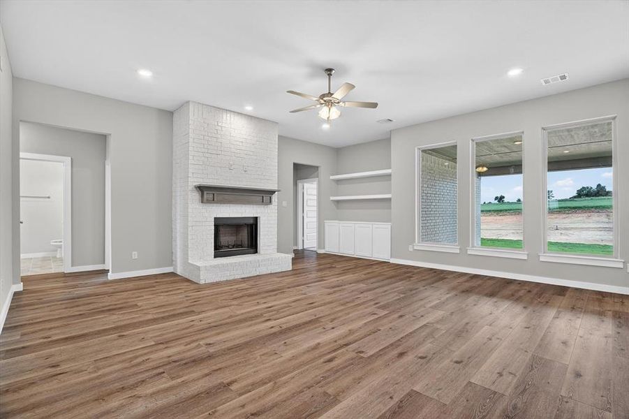 Spacious living area featuring wood-finish flooring, a white brick fireplace with a wood mantle, and recessed lighting Spacious living area featuring wood-finish flooring, a white brick fireplace with a wood mantle, and recessed lighting