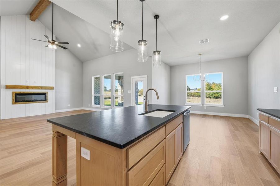 Kitchen with light brown cabinetry, light wood-style floors, an island with sink, a glass covered fireplace, and beamed ceiling
