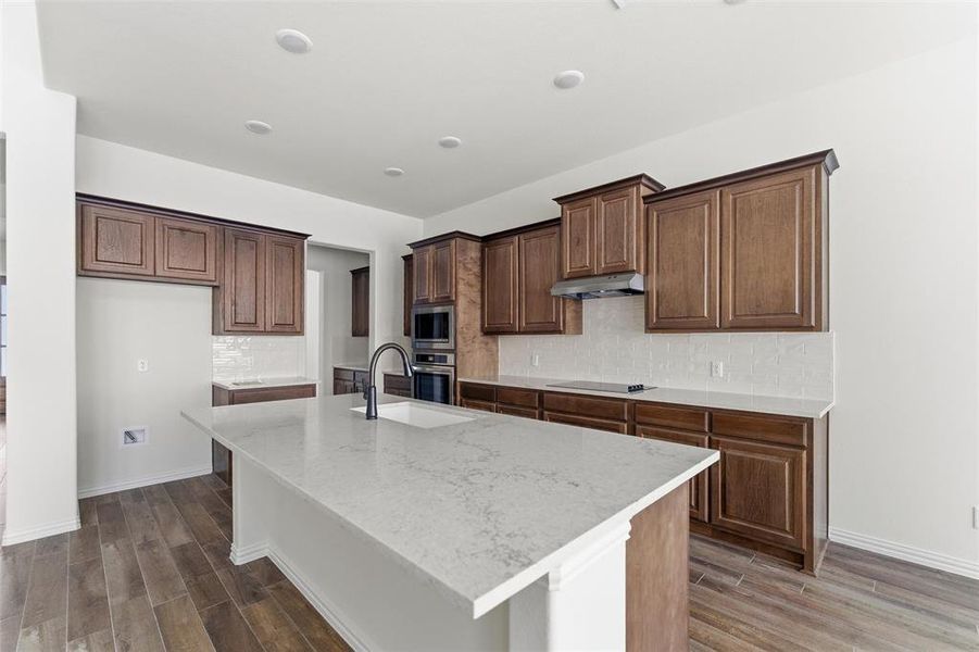 Kitchen with decorative backsplash, wood finish floors, a kitchen island with sink, light stone counters, and appliances with stainless steel finishes