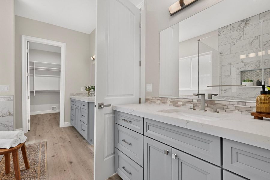 Double vanities with stone countertops stand on wide-plank wood flooring adjacent to a walk-in closet.