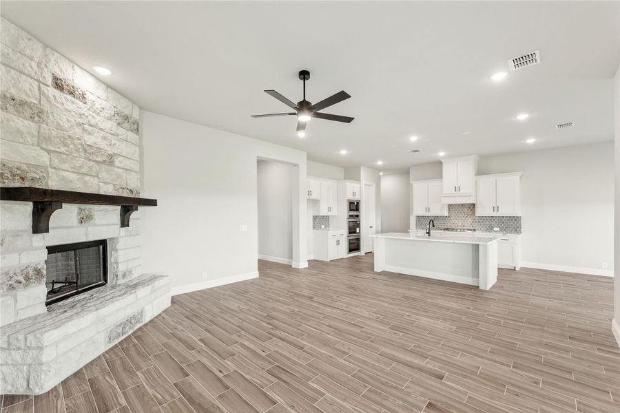 Open living area with stone fireplace, wood-look floors, and open kitchen with white cabinets and island in background Open living area with stone fireplace, wood-look floors, and open kitchen with white cabinets and island in background