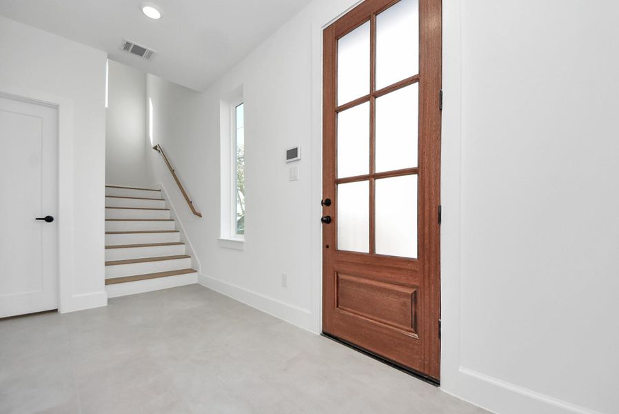 A bright, modern entryway featuring a wood front door with glass panels that allow natural light to flow throughout the space. Neutral walls, light tile flooring, and a staircase with a clean wood handrail set a polished, contemporary tone upon entry.