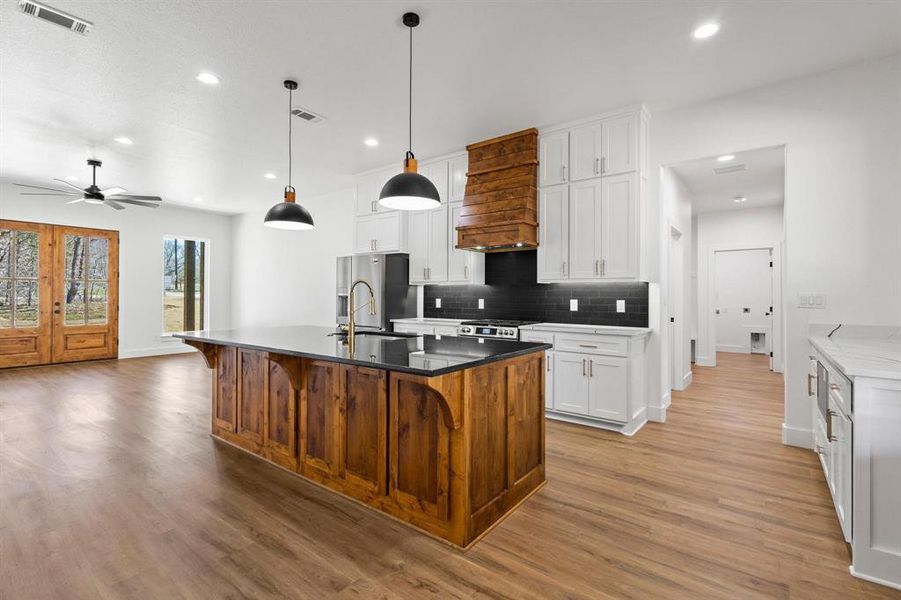 Kitchen with white cabinetry, a kitchen island with sink, decorative light fixtures, backsplash, and custom range hood