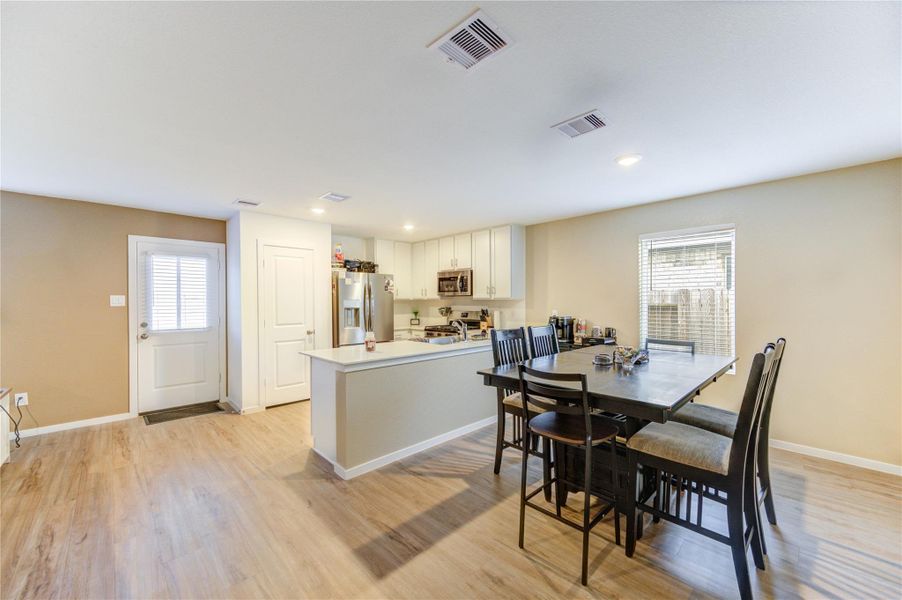 This photo showcases a bright, open-concept kitchen and dining area with modern appliances, white cabinetry, and vinyl plank flooring. The space features a large dining table, ample natural light from a window, and convenient access through a side door.