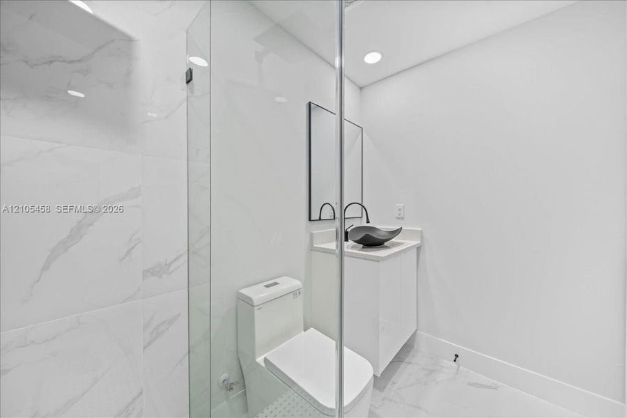 Glass-enclosed shower with large format tile surround, recessed lighting, modern vanity with vessel sink, and wall-mounted mirror Glass-enclosed shower with large format tile surround, recessed lighting, modern vanity with vessel sink, and wall-mounted mirror