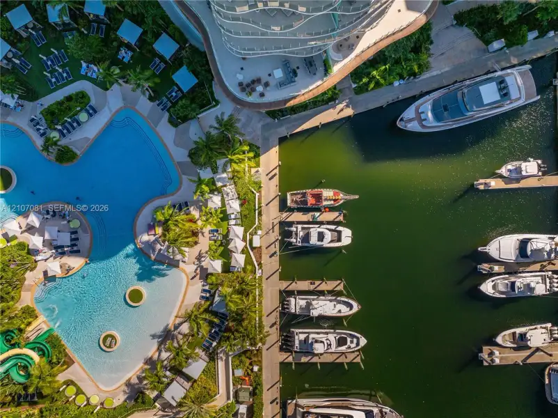 Aerial perspective of a multi-vessel deep-water marina featuring fixed concrete dockage and a large-scale swimming pool with integrated water slide and palm-landscaped deck Aerial perspective of a multi-vessel deep-water marina featuring fixed concrete dockage and a large-scale swimming pool with integrated water slide and palm-landscaped deck