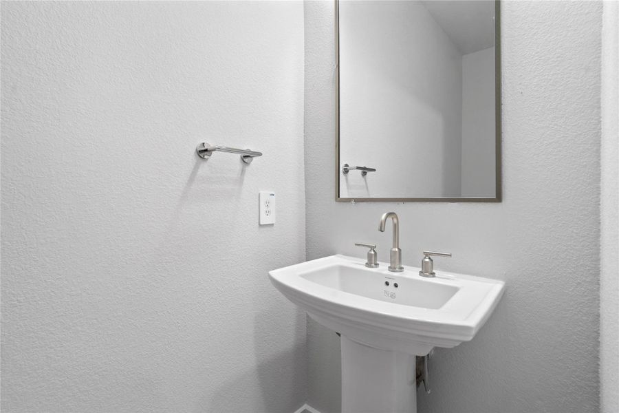 The first-floor powder room offers convenience and a modern aesthetic. It features a classic white pedestal sink complemented by a sleek faucet and a matching wall-mounted towel ring. A simple, large rectangular framed mirror enhances the sense of space and reflects the bright walls, making this half-bath both functional and stylish.