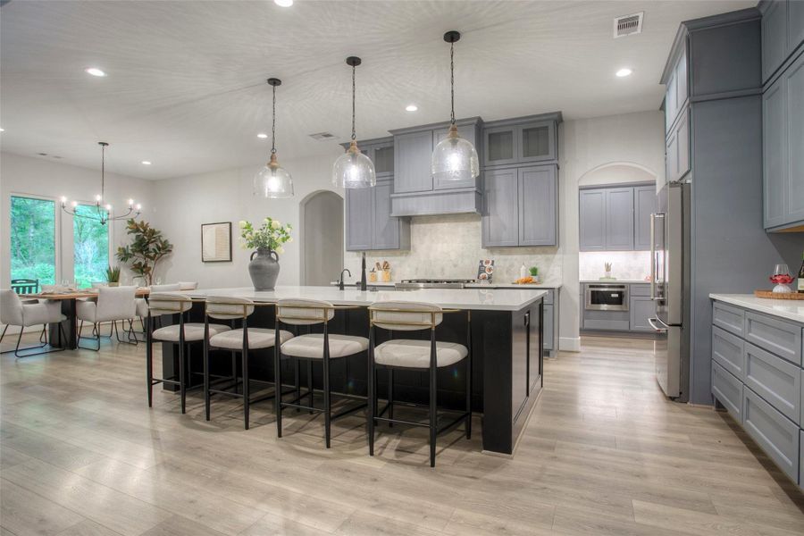 This spacious, modern kitchen features sleek gray cabinetry, a large island with seating, stainless steel appliances, and elegant pendant lighting. The open layout flows into a bright dining area with large windows overlooking greenery, creating a perfect space for entertaining.