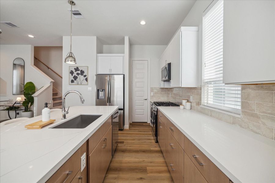 This sleek kitchen layout features a spacious quartz island with a modern stainless steel sink, perfectly complementing the stainless steel appliances. Ample storage is provided by the natural wood-tone cabinetry, while the neutral tile backsplash adds warmth and texture to the bright, open space.