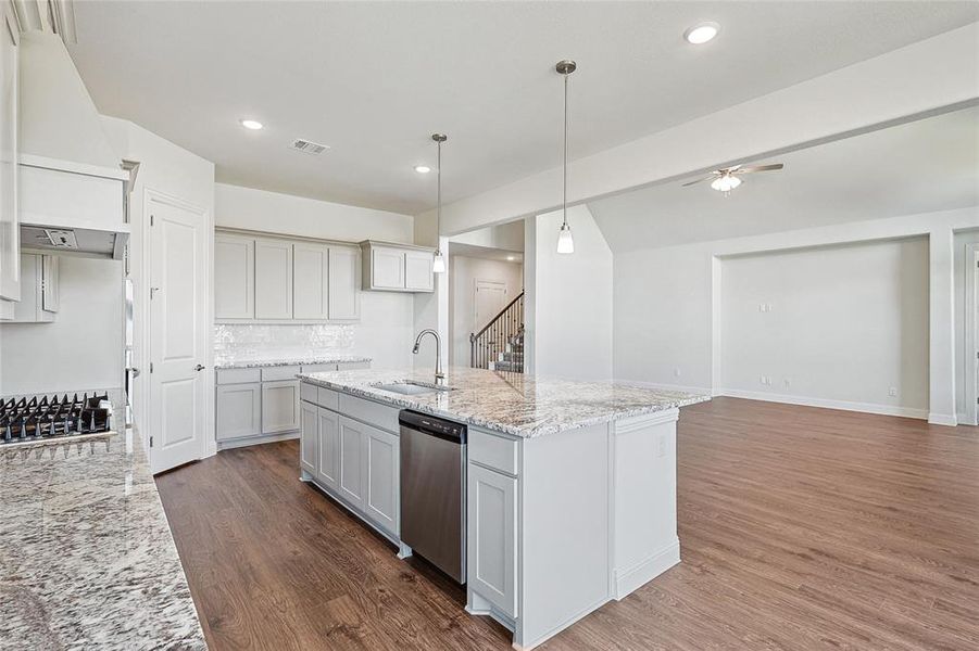 Kitchen with light stone counters, hanging light fixtures, a center island with sink, dark wood-type flooring, and dishwasher