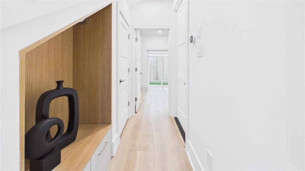 Bright, modern hallway with light wood flooring and white walls. Features a built-in niche with decorative art, leading to a sunlit glass door opening to a fenced outdoor area.
