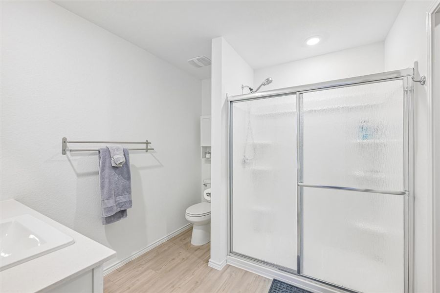 Bathroom featuring a glass-enclosed shower with sliding doors, a single vanity with an integrated sink, a toilet, and wood-finish flooring Bathroom featuring a glass-enclosed shower with sliding doors, a single vanity with an integrated sink, a toilet, and wood-finish flooring