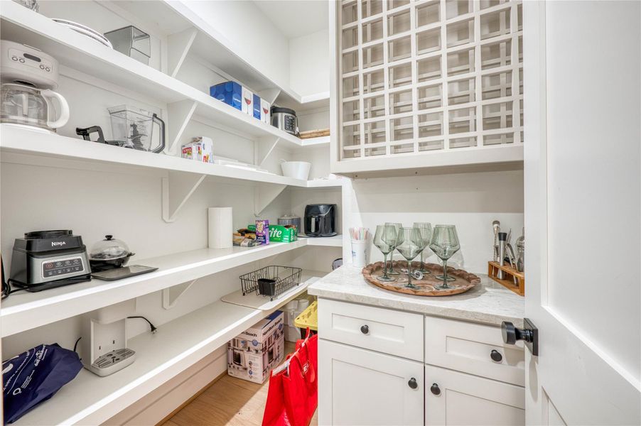 Walk-in pantry featuring extensive white shelving, integrated cabinetry with dark hardware, stone-finish countertops, and wood-finish flooring