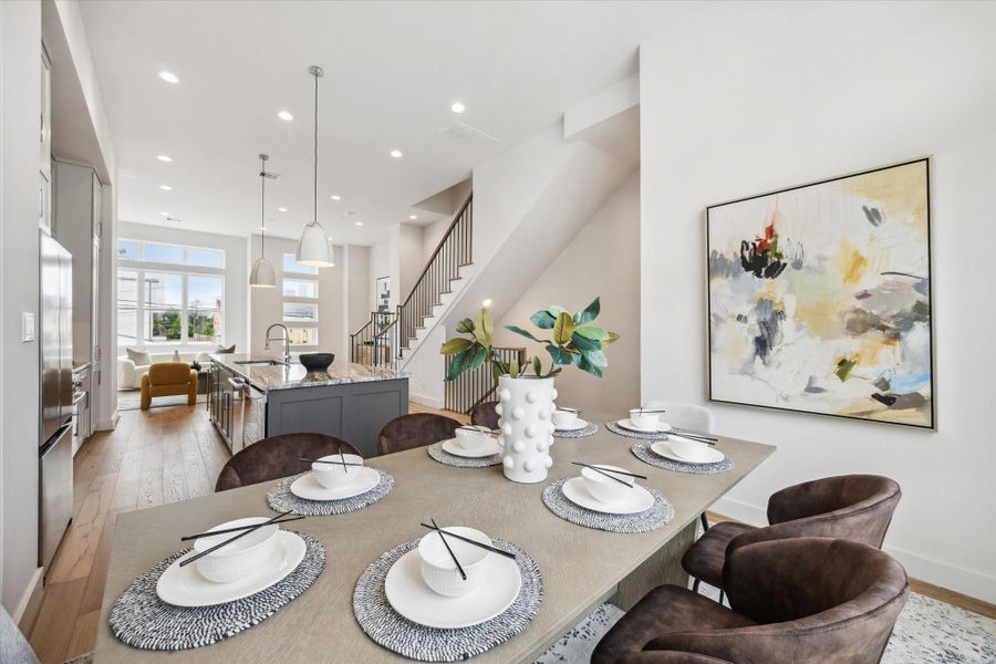 This photo showcases a modern, open-concept dining and kitchen area. It features a stylish dining table set for six, contemporary artwork, and a spacious kitchen with a large island. Natural light floods in through large windows, and there's a view of a cozy living area. The design is sleek with neutral tones and elegant finishes.