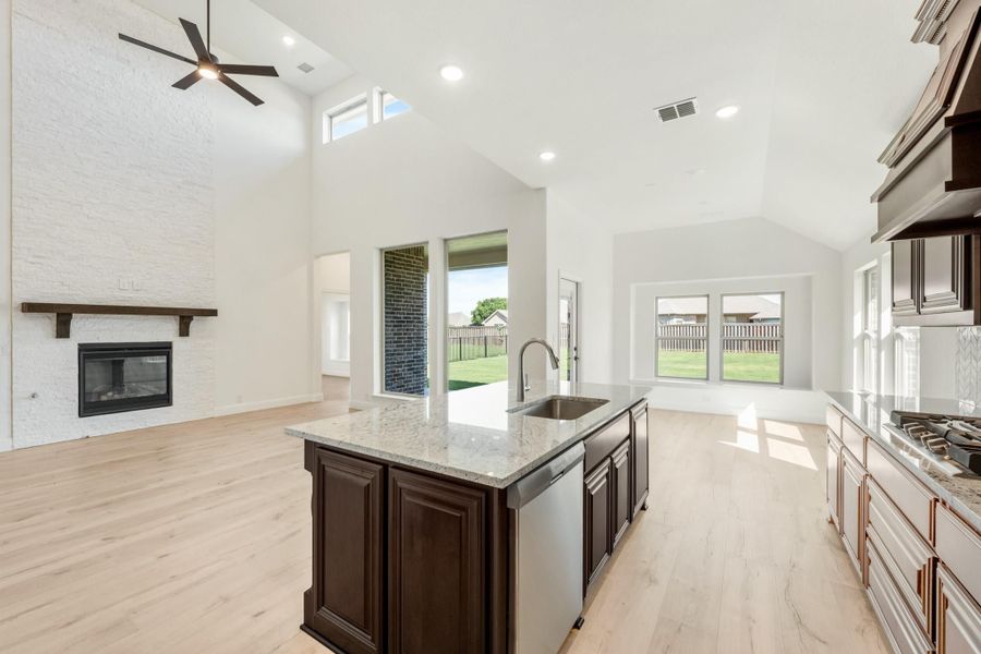 Open-concept kitchen with granite island, dark cabinets, and vaulted ceiling adjoining living room with brick fireplace
