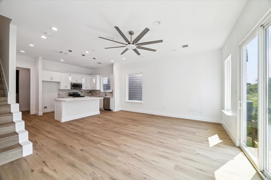 From this angle, the open layout between the living area and kitchen truly shines. A spacious footprint allows plenty of room for comfortable seating and dining arrangements, while the large ceiling fan adds style and everyday comfort. Natural light pours in through expansive windows and glass doors, highlighting the warm flooring and crisp neutral walls. The kitchen remains visually connected for easy conversation and entertaining.