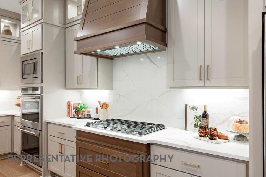 Kitchen featuring premium range hood, appliances with stainless steel finishes, tasteful backsplash, glass insert cabinets, and light stone counters Kitchen featuring premium range hood, appliances with stainless steel finishes, tasteful backsplash, glass insert cabinets, and light stone counters