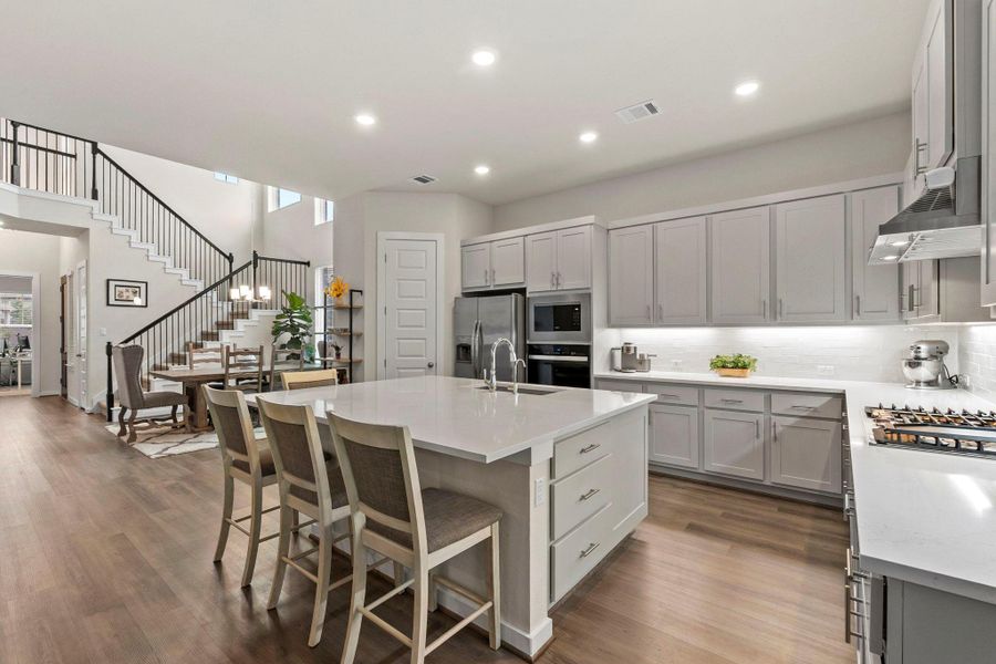 Spacious kitchen with a large central island, light wood-finish flooring, recessed lighting, stainless steel appliances, and ample grey cabinetry with undermount lighting