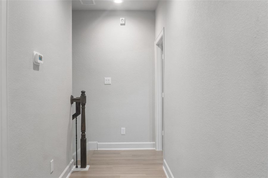 This photo shows a clean, narrow hallway with light gray walls and hardwood floors. It features a wooden railing leading to a staircase, a thermostat, and recessed lighting, creating a simple and modern look.