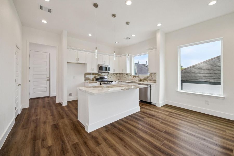Clean, bright, and beautifully finished, this open kitchen features white shaker cabinetry, striking stone-look surfaces, stainless steel appliances, and a large center island. Three pendant lights add a sleek decorative touch above the island, while recessed lighting provides excellent illumination throughout. Warm wood-look flooring softens the crisp palette and creates a seamless transition into the adjacent dining and living spaces. Large windows add natural light and enhance the open, airy feel Clean, bright, and beautifully finished, this open kitchen features white shaker cabinetry, striking stone-look surfaces, stainless steel appliances, and a large center island. Three pendant lights add a sleek decorative touch above the island, while recessed lighting provides excellent illumination throughout. Warm wood-look flooring softens the crisp palette and creates a seamless transition into the adjacent dining and living spaces. Large windows add natural light and enhance the open, airy feel
