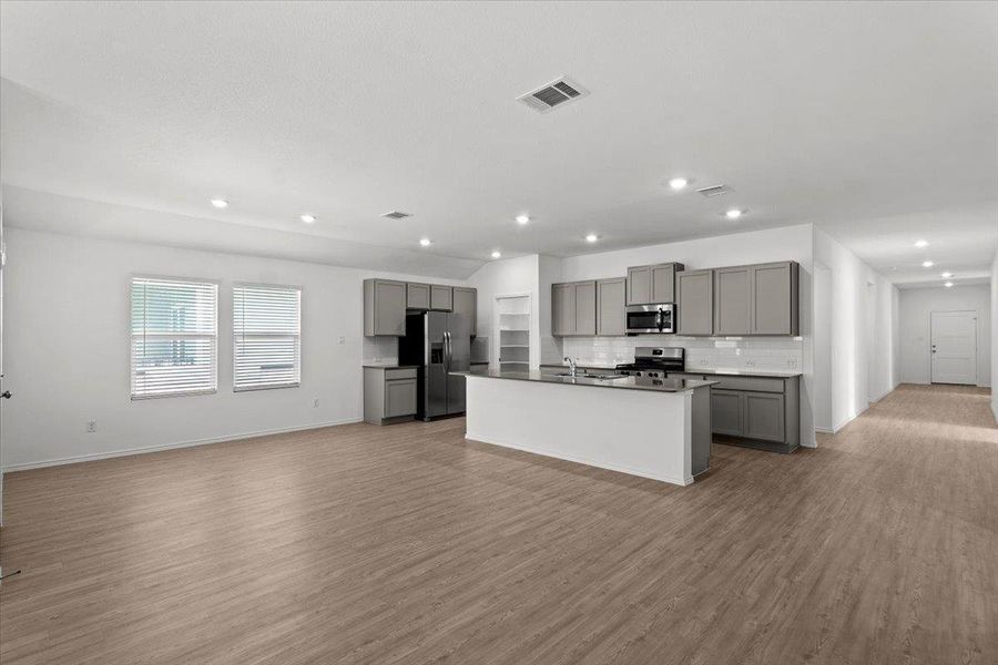 An open-concept layout unites the kitchen and living area, featuring an island with a sink, sleek gray cabinetry, and light wood flooring. An open-concept layout unites the kitchen and living area, featuring an island with a sink, sleek gray cabinetry, and light wood flooring.
