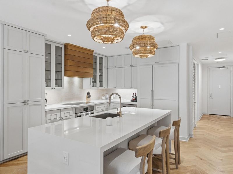 Modern kitchen featuring a large center island with a sink, light-toned cabinetry, integrated appliances, and wood-finish herringbone flooring Modern kitchen featuring a large center island with a sink, light-toned cabinetry, integrated appliances, and wood-finish herringbone flooring
