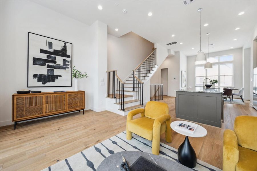 This modern living space features an open-concept design with light wood flooring, a stylish kitchen island, and a cozy seating area with vibrant yellow chairs. Large windows provide ample natural light, and a sleek staircase adds a contemporary touch.