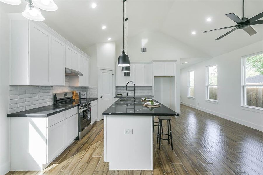 This is a spacious, modern kitchen featuring white cabinetry, dark countertops, stainless steel appliances, and a central island with a sink. The room has hardwood floors, recessed lighting, a vaulted ceiling, and ample natural light from several windows. A stylish pendant light hangs over the island.