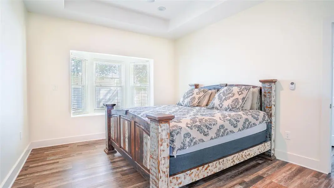 Primary bedroom with wood-style floors, bay window with blinds, and a raised ceiling Primary bedroom with wood-style floors, bay window with blinds, and a raised ceiling