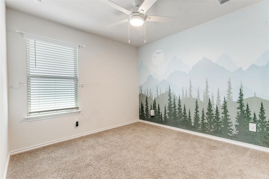 Carpeted room featuring a white ceiling fan with integrated lighting, a single window with blinds, and a decorative wall mural depicting mountains and trees Carpeted room featuring a white ceiling fan with integrated lighting, a single window with blinds, and a decorative wall mural depicting mountains and trees