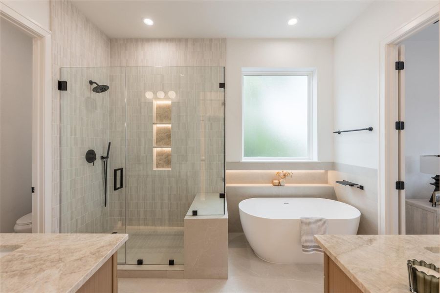 An en suite that treats the bath like a room, not a utility. Vertical stacked tile climbs the shower wall, a backlit stone niche glows at eye level, a freestanding soaker sits under a privacy window with a stone sill, and twin vanities bookend the space in veined quartzite. Matte black fixtures carry the hardware story throughout.