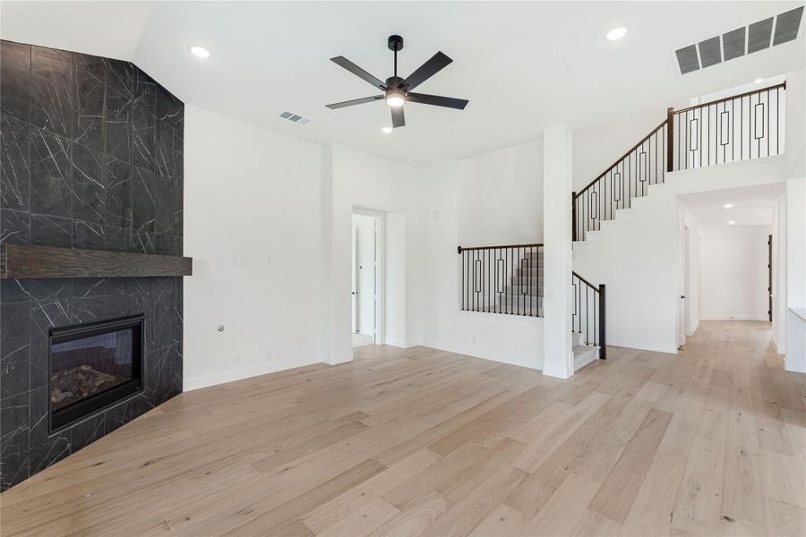 Open living room with dark stone fireplace, light hardwood floors, ceiling fan, and staircase with metal railings