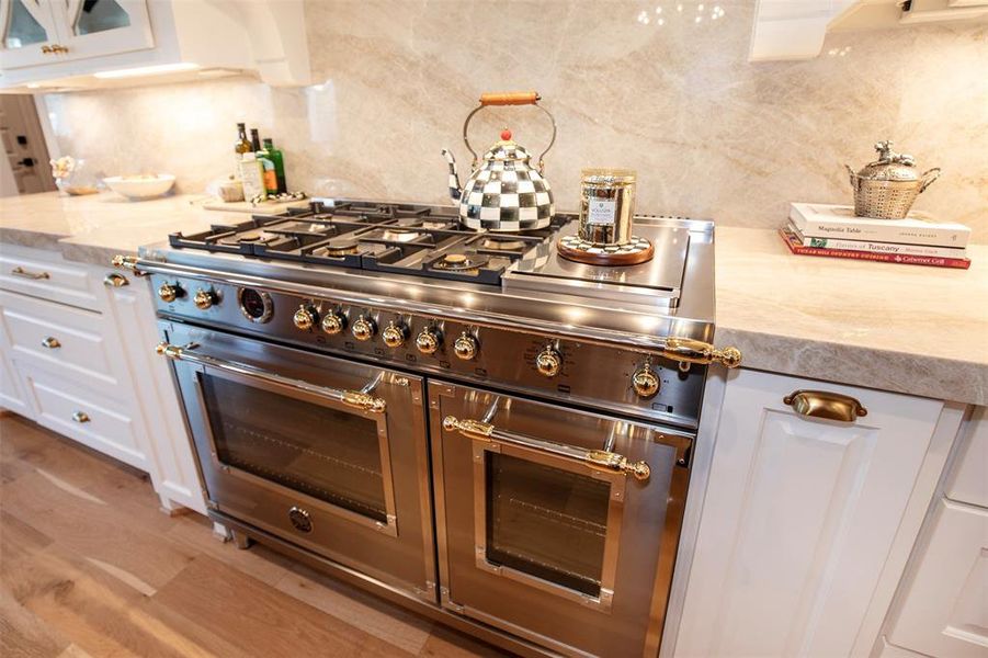 This kitchen features a luxurious stainless steel range with gold accents, set against a sleek, marble backsplash. The surrounding cabinetry is white with elegant gold handles, and the wooden floor adds warmth to the space.