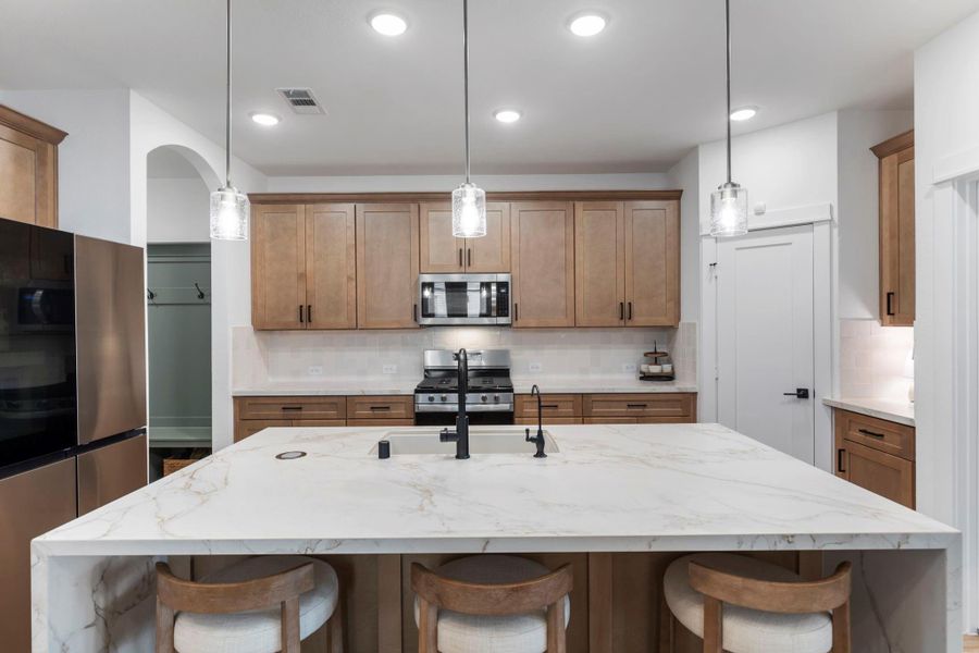 This modern kitchen features a spacious island with seating, sleek pendant lighting, and stainless steel appliances. The light wood cabinetry offers ample storage, complementing the room's bright and contemporary design.