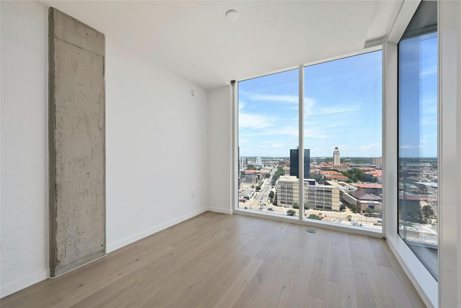 Step into this sun-drenched bedroom featuring expansive floor-to-ceiling windows that offer breathtaking panoramic views of the city skyline. The clean, modern design is highlighted by light hardwood flooring, crisp white walls, and a striking exposed concrete column that adds a touch of industrial elegance.