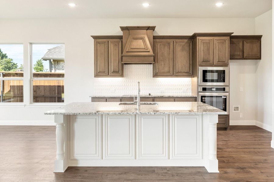 Kitchen with white island, granite countertops, brown cabinets, decorative range hood, and stainless steel appliances