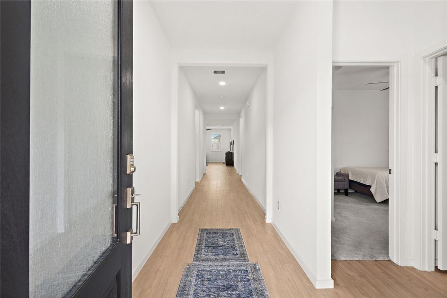 A bright, welcoming hallway with light wood flooring and white walls. It features a long runner rug and leads to multiple rooms, including a visible bedroom on the right. The space is well-lit with recessed lighting.