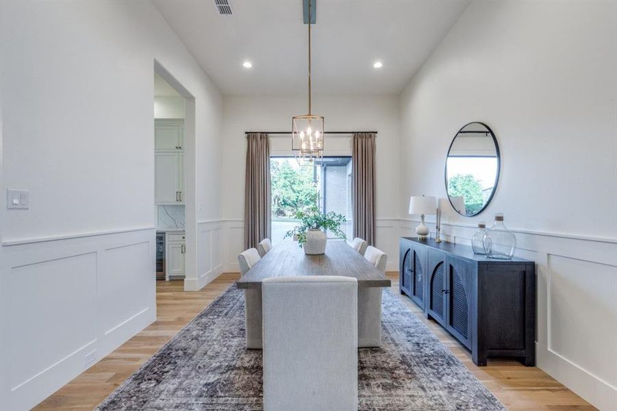 Dining area featuring a wainscoted wall, a decorative wall, light wood-type flooring, a chandelier, and wine cooler Dining area featuring a wainscoted wall, a decorative wall, light wood-type flooring, a chandelier, and wine cooler