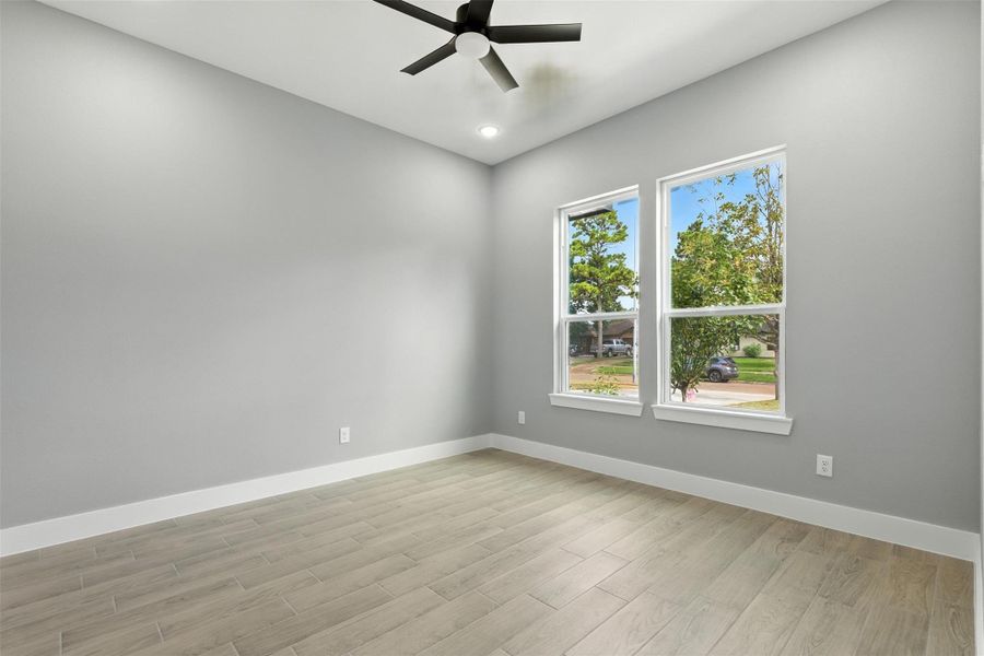 Bright, modern room with light flooring, gray walls, and a ceiling fan. Features two large windows providing ample natural light and a view of outside.