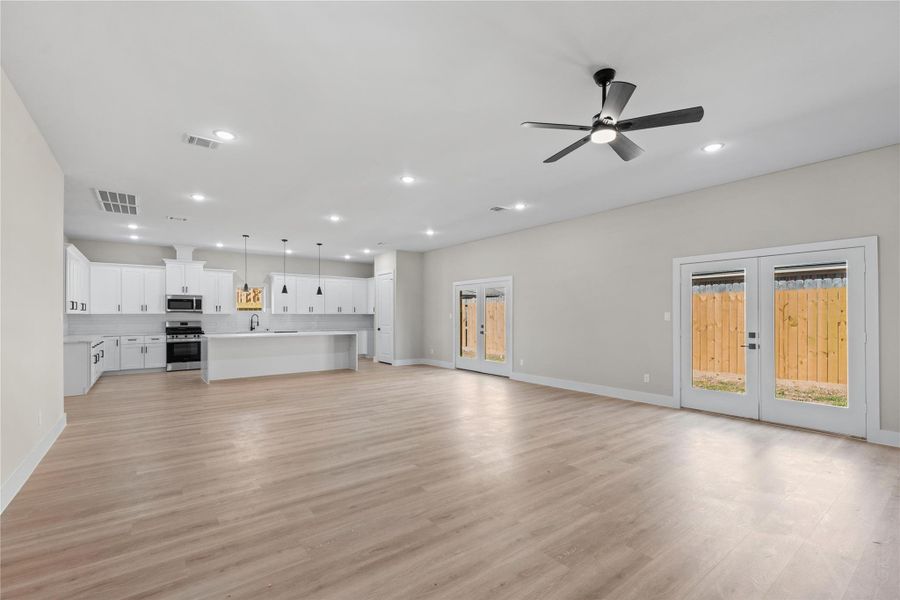 Spacious open-concept living area with light wood flooring, a modern kitchen featuring white cabinetry and stainless steel appliances, a large island with pendant lighting, a ceiling fan, and double French doors leading to a fenced backyard.