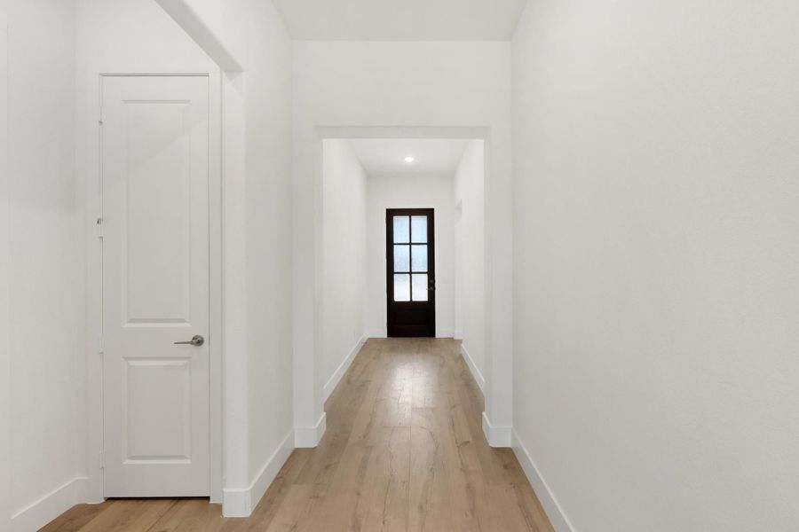 Hallway with white walls, light wood floors, and dark glass-panel front door at the end Hallway with white walls, light wood floors, and dark glass-panel front door at the end